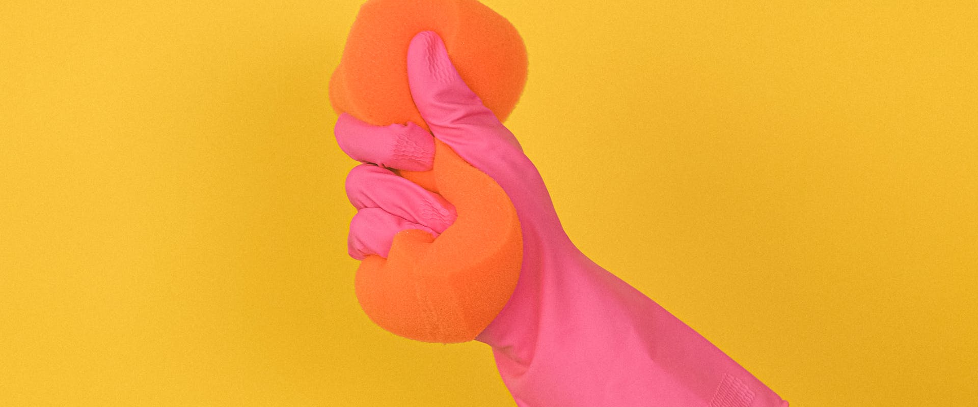 Pink rubber-glove hand squeezing an orange sponge on a sunny yellow background — Airbnb turnover cleaning
