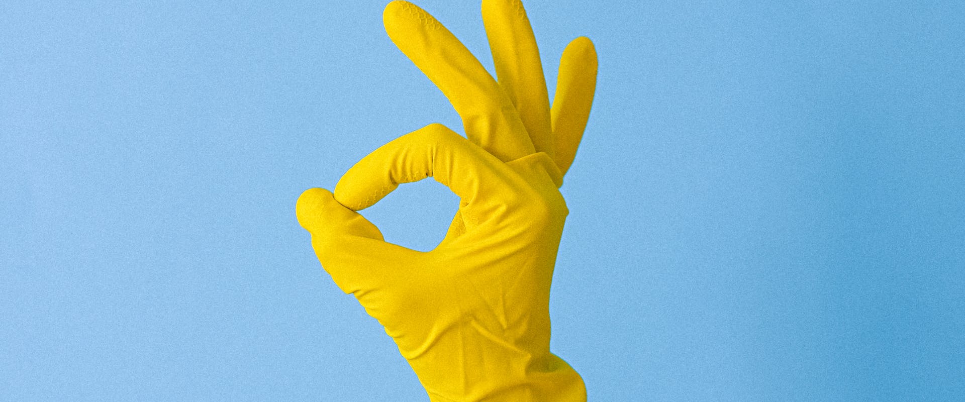 Yellow rubber-glove hand making the OK gesture on a powder-blue background — symbolising professional commercial cleaning